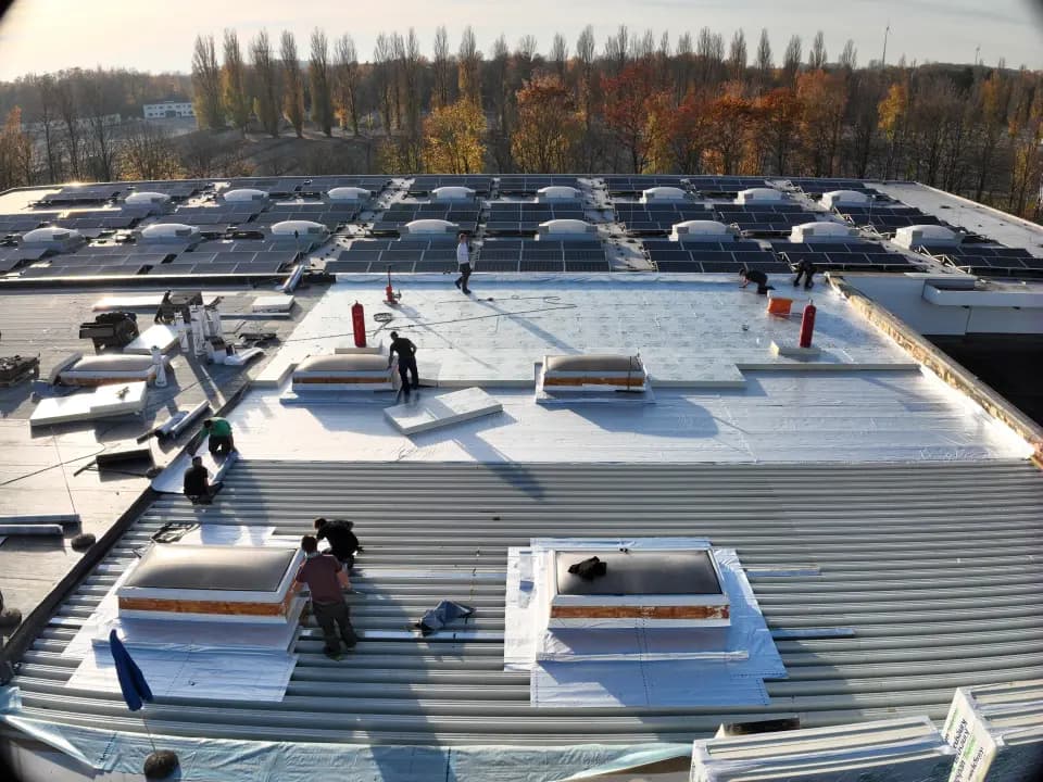 Drohnenaufnahme einer Flachdach-Baustelle mit PV-Anlage und Dachdeckern bei der Arbeit — Baufortschritt aus der Luft dokumentiert
